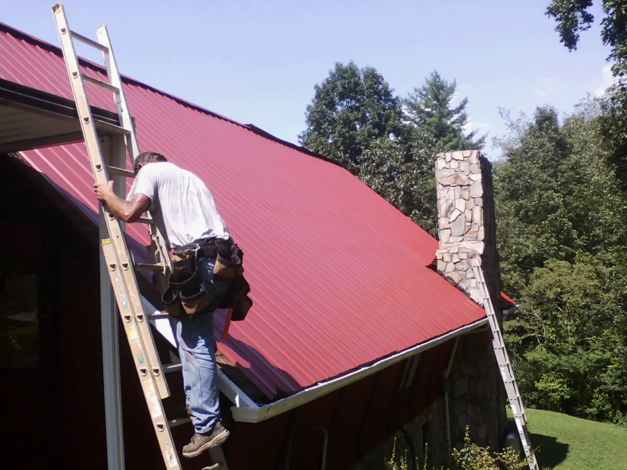 Roofer on a ladder beside a red metal roof and stone chimney — Soffit & Fascia Repair by Roofing Brothers in Claysburg